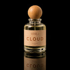 A clear glass perfume bottle with a round wooden stopper and a beige label reading 'ATHRISE BLOOM', centered on a black reflective background.