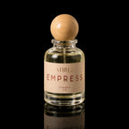 A clear glass perfume bottle with a light wood spherical cap and a beige label reading 'ATHRI EMPRESS EAU DE PARFUM 50 ML', photographed on a black reflective surface.