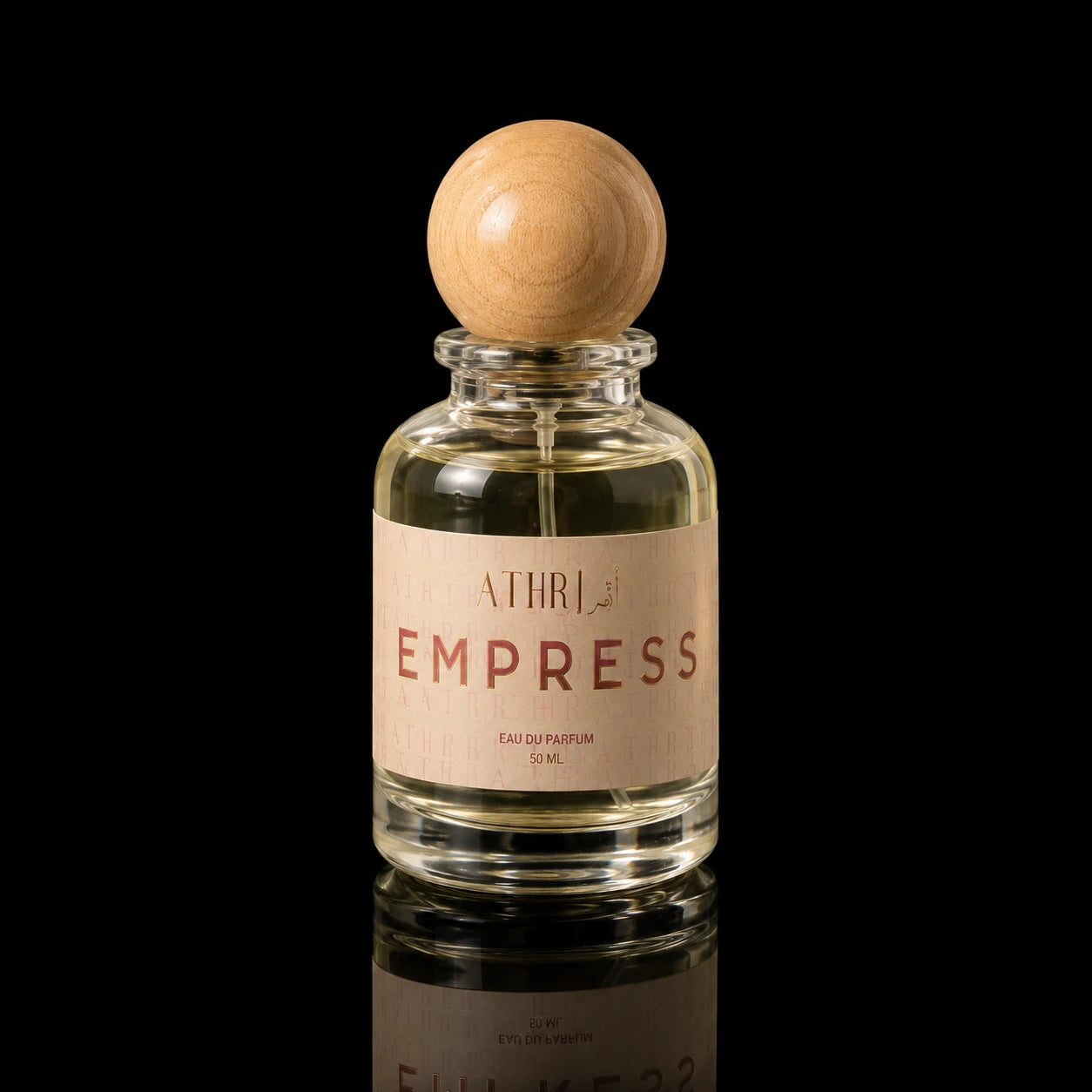 A clear glass perfume bottle with a light wood spherical cap and a beige label reading 'ATHRI EMPRESS EAU DE PARFUM 50 ML', photographed on a black reflective surface.