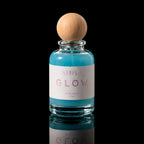 A clear glass perfume bottle filled with bright turquoise-blue liquid, featuring a white label with the text 'ATIRL' and 'GLOW' in pink and gold lettering, topped with a round natural wood cap, set against a solid black background.
