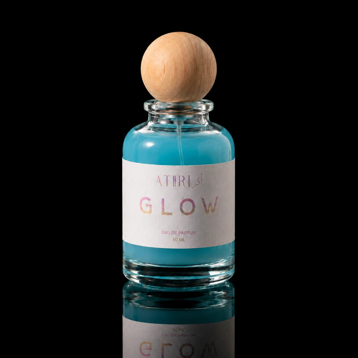A clear glass perfume bottle filled with bright turquoise-blue liquid, featuring a white label with the text 'ATIRL' and 'GLOW' in pink and gold lettering, topped with a round natural wood cap, set against a solid black background.