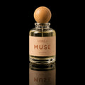 A clear glass perfume bottle with a round light wood cap and a beige label reading 'ATHLE MUSE', photographed against a black background with a reflection below.