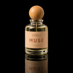 A clear glass perfume bottle with a round light wood cap and a beige label reading 'ATHLE MUSE', photographed against a black background with a reflection below.