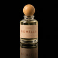 A clear glass perfume bottle with a round light wood cap and a beige label reading ATHRI ROMELLA, photographed against a black background with a reflection below.
