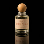 A clear glass perfume bottle with a round light wood cap and a beige label reading ATHRI ROMELLA, photographed against a black background with a reflection below.