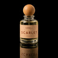 A clear glass perfume bottle with a round wooden cap and a tan paper label reading 'ATHRIE SCARLET EAU DE PARFUM'. The bottle is centered on a reflective black surface against a black background.