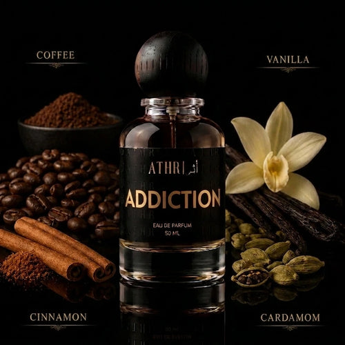A 50 ml bottle of ATHRI Addiction Eau de Parfum centered against a black background, surrounded by its scent ingredients: coffee beans, cinnamon sticks, vanilla flower, cardamom pods, and coffee powder. The bottle has a black label with gold lettering and a black wooden dropper cap.