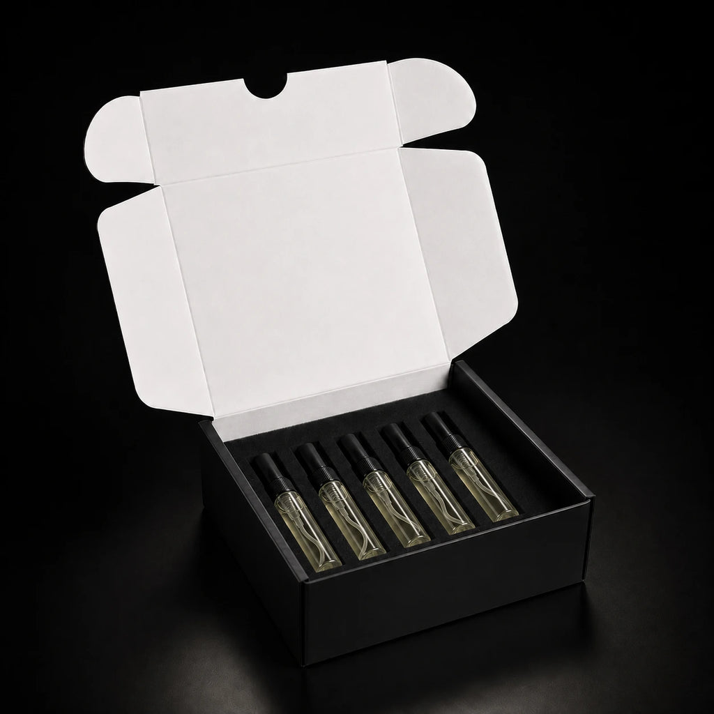 An open black gift box with a white inner flap, holding five small clear glass perfume or fragrance bottles with black caps on a black background.