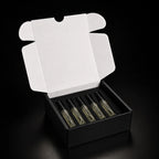 An open black gift box with a white inner flap, holding five small clear glass perfume or fragrance bottles with black caps on a black background.