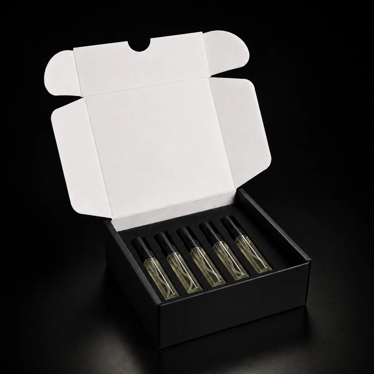 An open black gift box with a white inner flap, holding five small clear glass perfume or fragrance bottles with black caps on a black background.