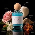 A clear glass perfume bottle filled with bright aqua blue liquid, topped with a round natural wood cap, centered on a black reflective surface. The bottle is surrounded by fragrance-themed props and labeled ingredients: a pink rose, white jasmine flowers, a fluffy white pom-pom labeled MUSK, a pile of beige powder labeled POWDER, and amber crystals labeled AMBER.