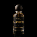 A 50 ml bottle of Athri Mango Rush Eau de Parfum, featuring a clear glass bottle filled with amber-colored fragrance liquid, a black label with gold lettering, and a dark wooden spherical cap, set against a solid black background with a subtle reflection below.