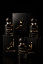 Four perfume bottles and matching black boxes arranged on a black studio background. The bottles are clear glass with amber-brown liquid and dark round caps, labeled with gold text including ATHRI, TROPICA, and COCOVERA.