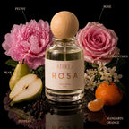 A clear glass perfume bottle with a round light wood cap stands centered on a reflective black surface, surrounded by fragrance-themed props including pink flowers, a pear, mandarin orange, peppercorns, and small white blossoms. Text labels point to the scent notes: Peony, Rose, Pear, Osmanthus, Mandarin Orange, and Peppers.