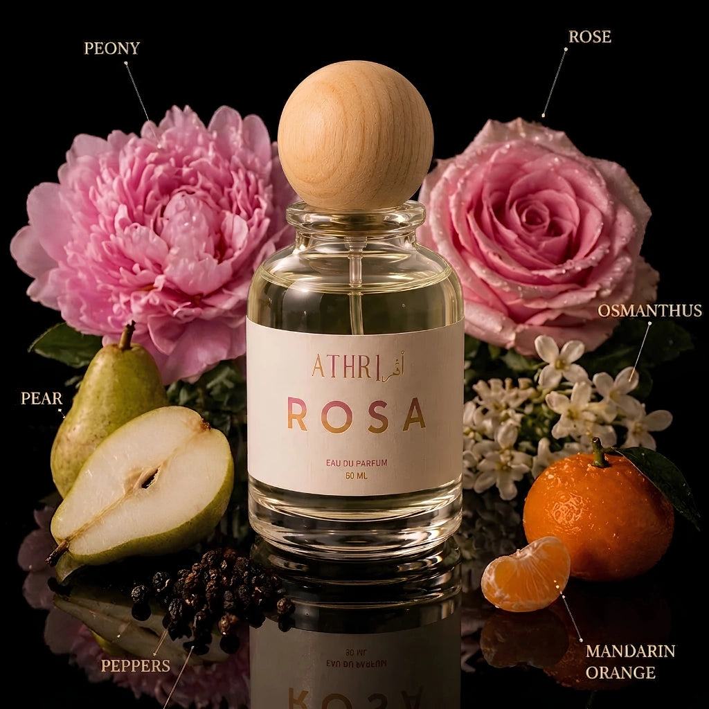 A clear glass perfume bottle with a round light wood cap stands centered on a reflective black surface, surrounded by fragrance-themed props including pink flowers, a pear, mandarin orange, peppercorns, and small white blossoms. Text labels point to the scent notes: Peony, Rose, Pear, Osmanthus, Mandarin Orange, and Peppers.