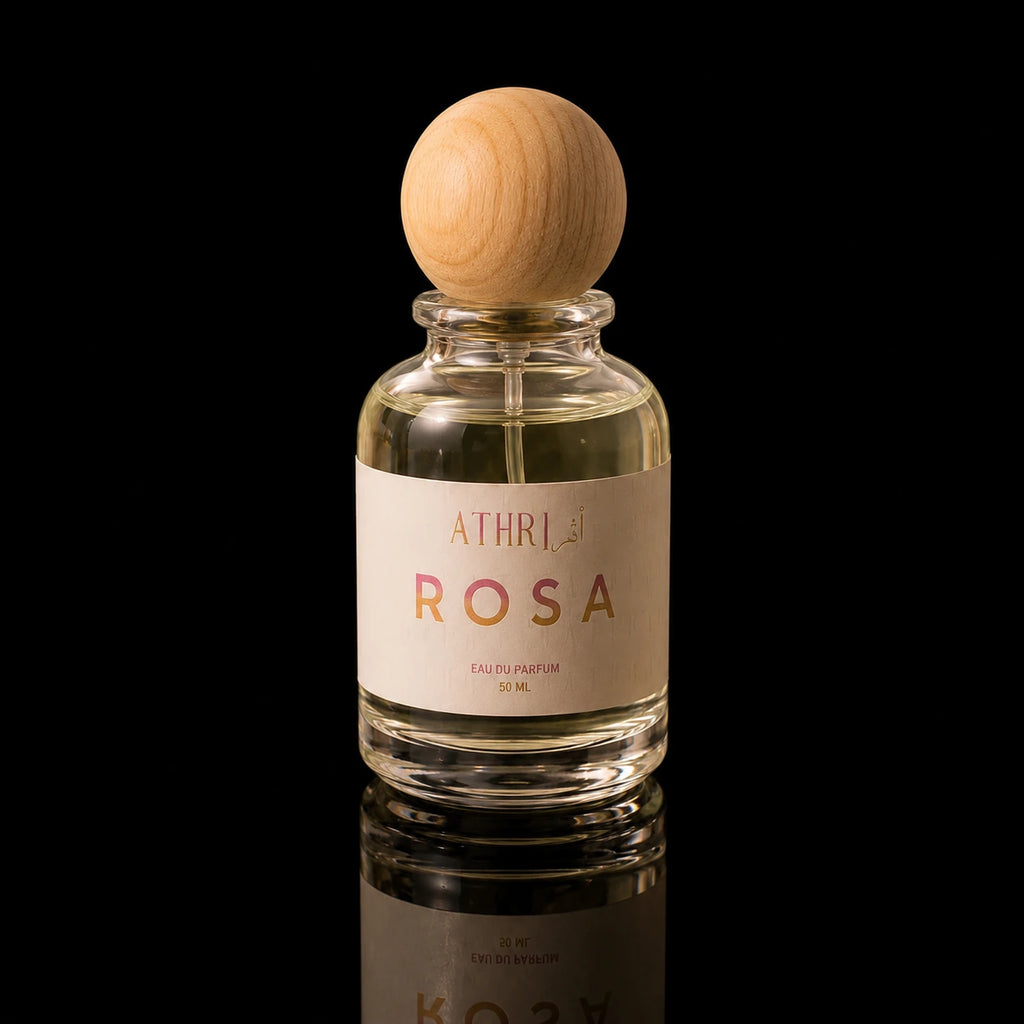 A clear glass perfume bottle with a light wood spherical cap, filled with pale yellow liquid. The bottle has a cream-colored label with the brand name 'ATHRI' and the scent name 'ROSA' in pink lettering.