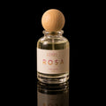 A clear glass perfume bottle with a light wood spherical cap, filled with pale yellow liquid. The bottle has a cream-colored label with the brand name 'ATHRI' and the scent name 'ROSA' in pink lettering.