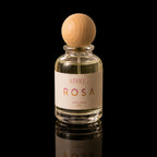 A clear glass perfume bottle with a light wood spherical cap, filled with pale yellow liquid. The bottle has a cream-colored label with the brand name 'ATHRI' and the scent name 'ROSA' in pink lettering.